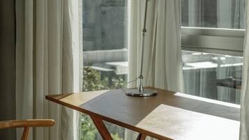 Una habitación con un escritorio y silla de madera, una lámpara de pie y vistas a edificios a través de cortinas traslúcidas.