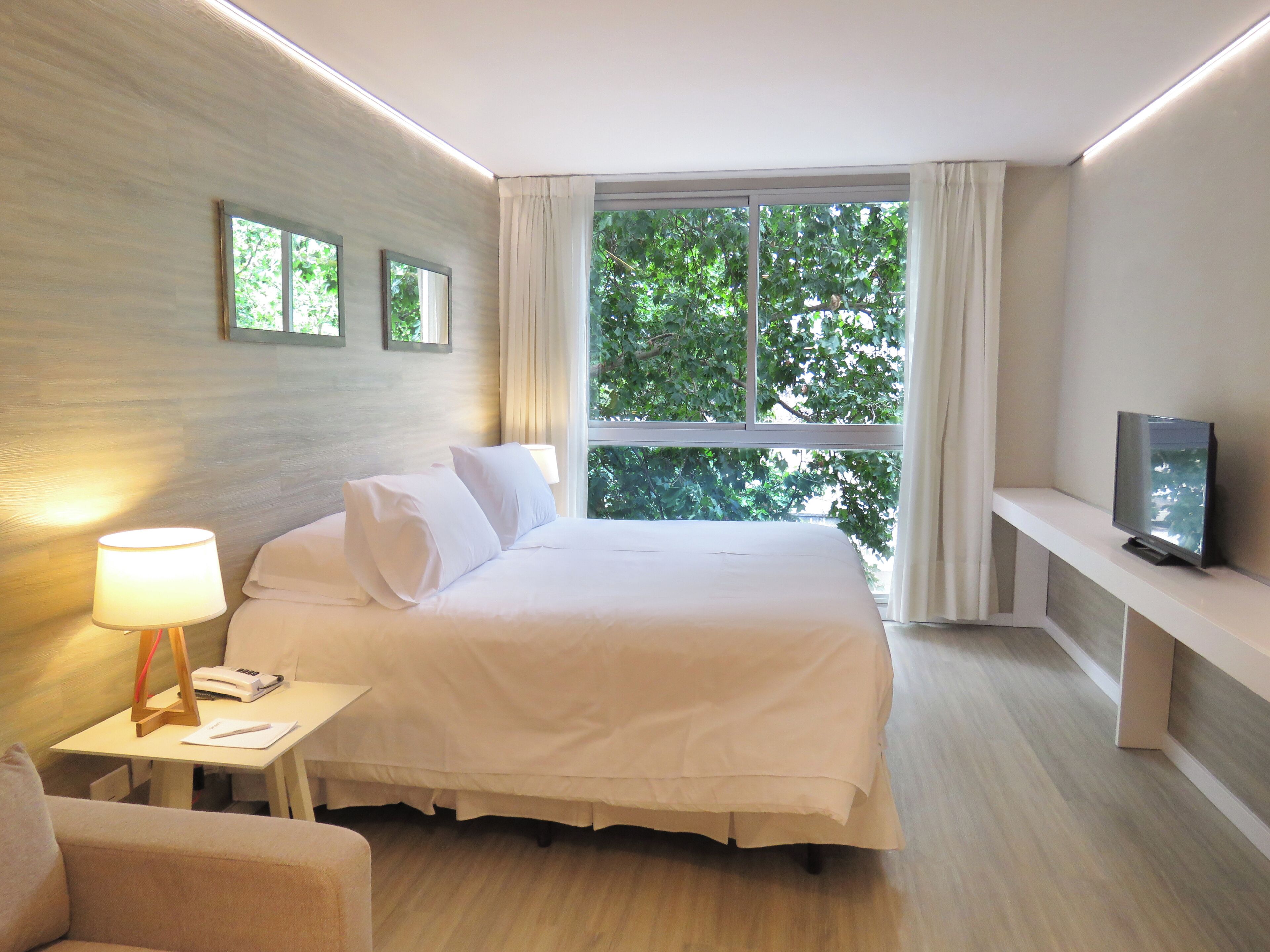 A modern hotel room with a large bed, a bedside table with a lamp, a TV, and a window with curtains.