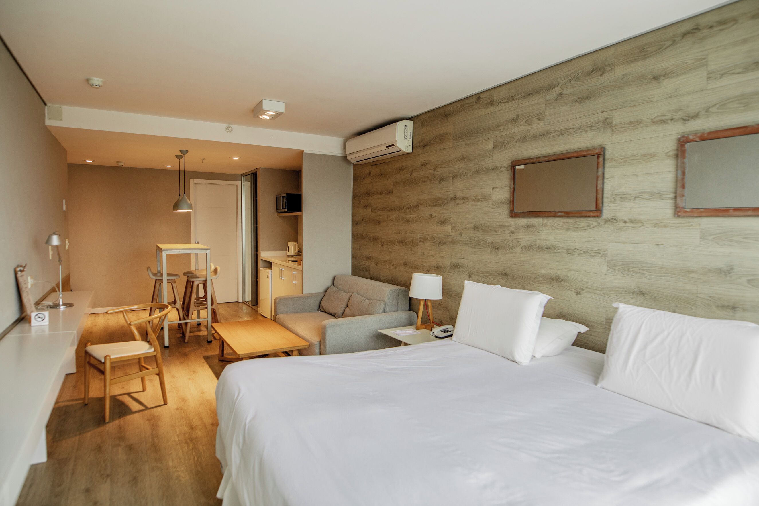 Een hotelkamer met een groot bed, een bureau, een stoel, een kleine tafel en een bank. Er is een airconditioningunit aan de muur en een deur die leidt naar een andere kamer.