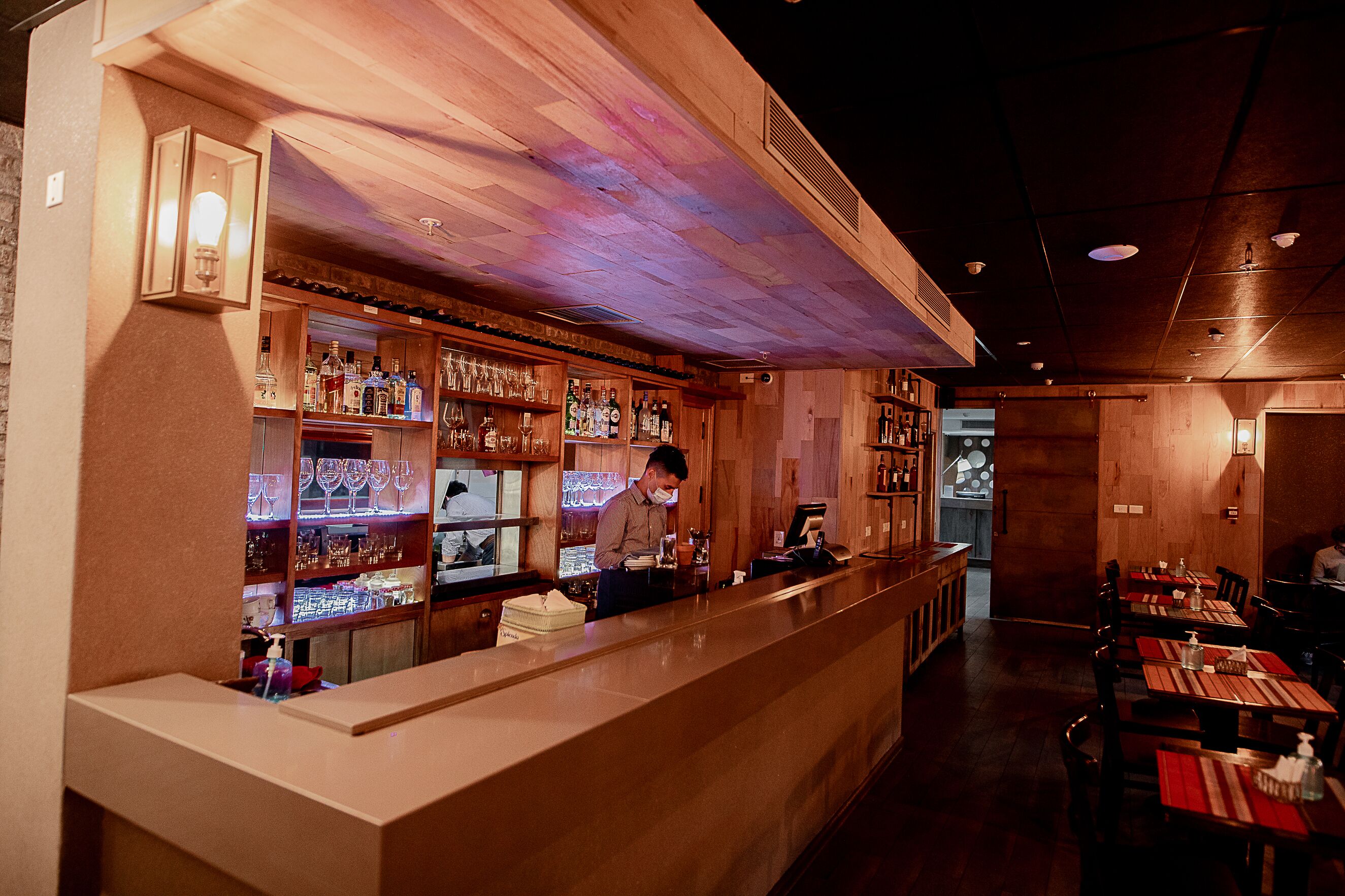 A bar with wooden shelves stocked with bottles and glasses, a counter with a barista, and tables with red placemats in a dimly lit interior.
