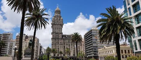 A city square with a prominent building featuring a tower, surrounded by palm trees and a fountain.