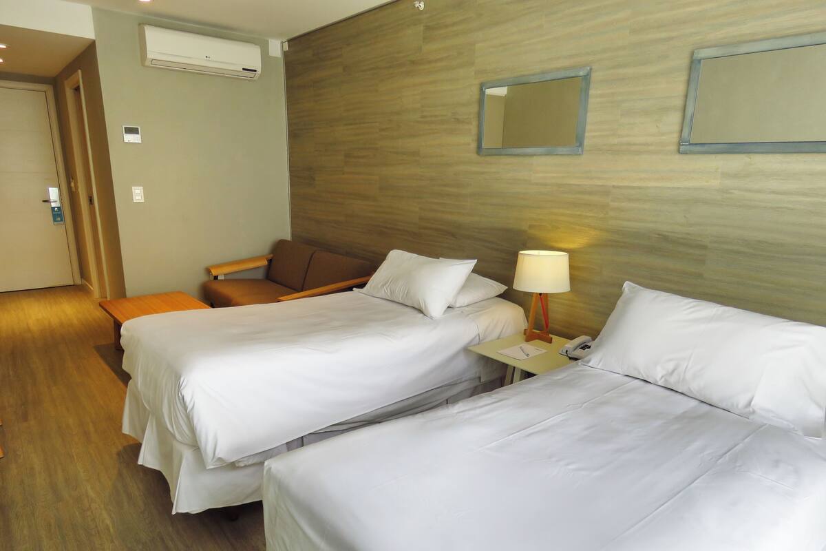Quarto de hotel com duas camas, uma mesa pequena com abajur e cabeceira de madeira.