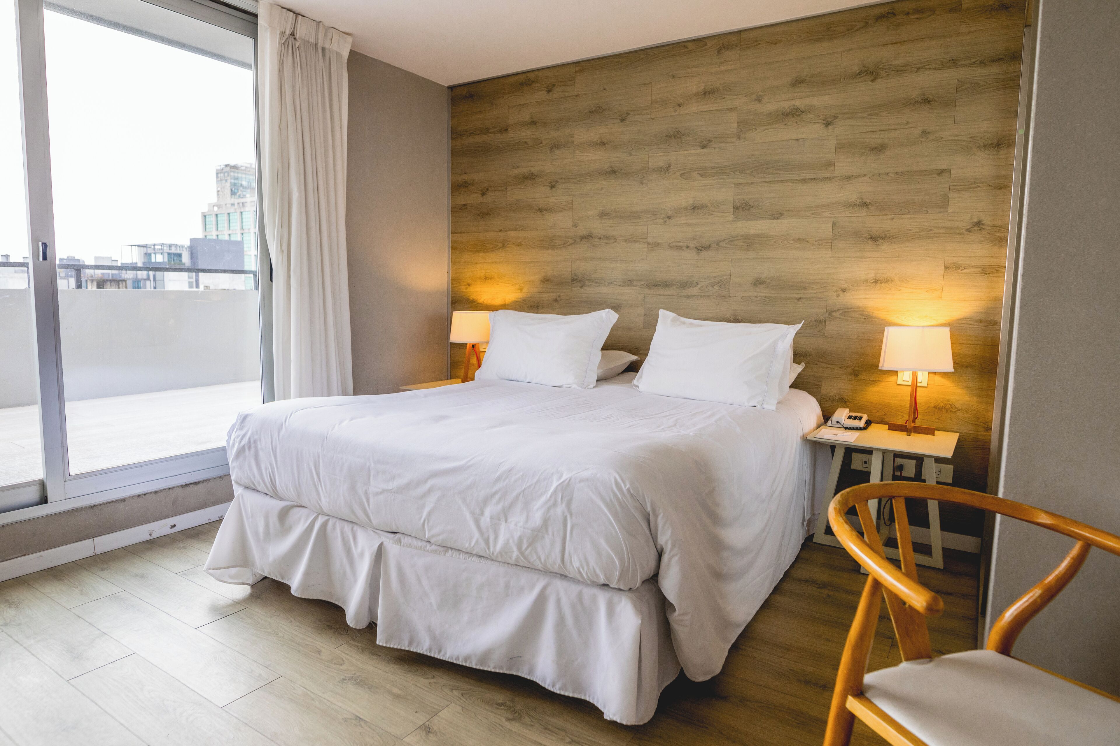 A hotel room with a large bed, two bedside lamps, a wooden headboard, a wooden chair, and a view of the city through a large window.