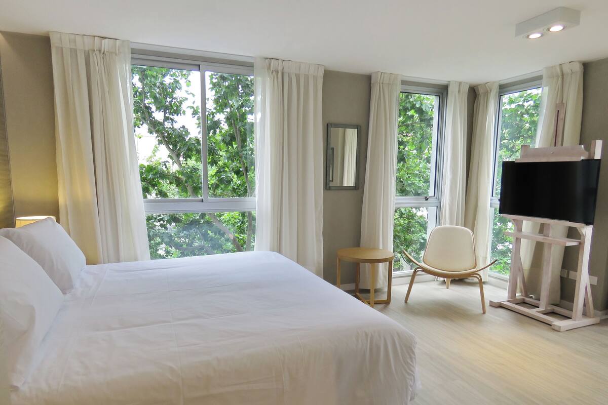 Quarto de hotel com uma cama grande, uma cadeira, uma TV e janelas amplas com cortinas.