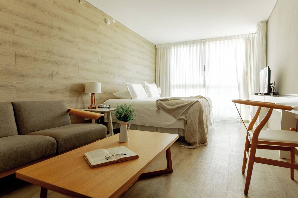 Quarto de hotel moderno com piso de madeira, cama com lençóis brancos, um sofá, uma escrivaninha com luminária e uma televisão.