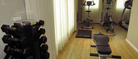 Ein Heimfitnessraum mit Trainingsgeräten, darunter Kurzhanteln, eine Bank, ein Ergometer und ein Crosstrainer.
