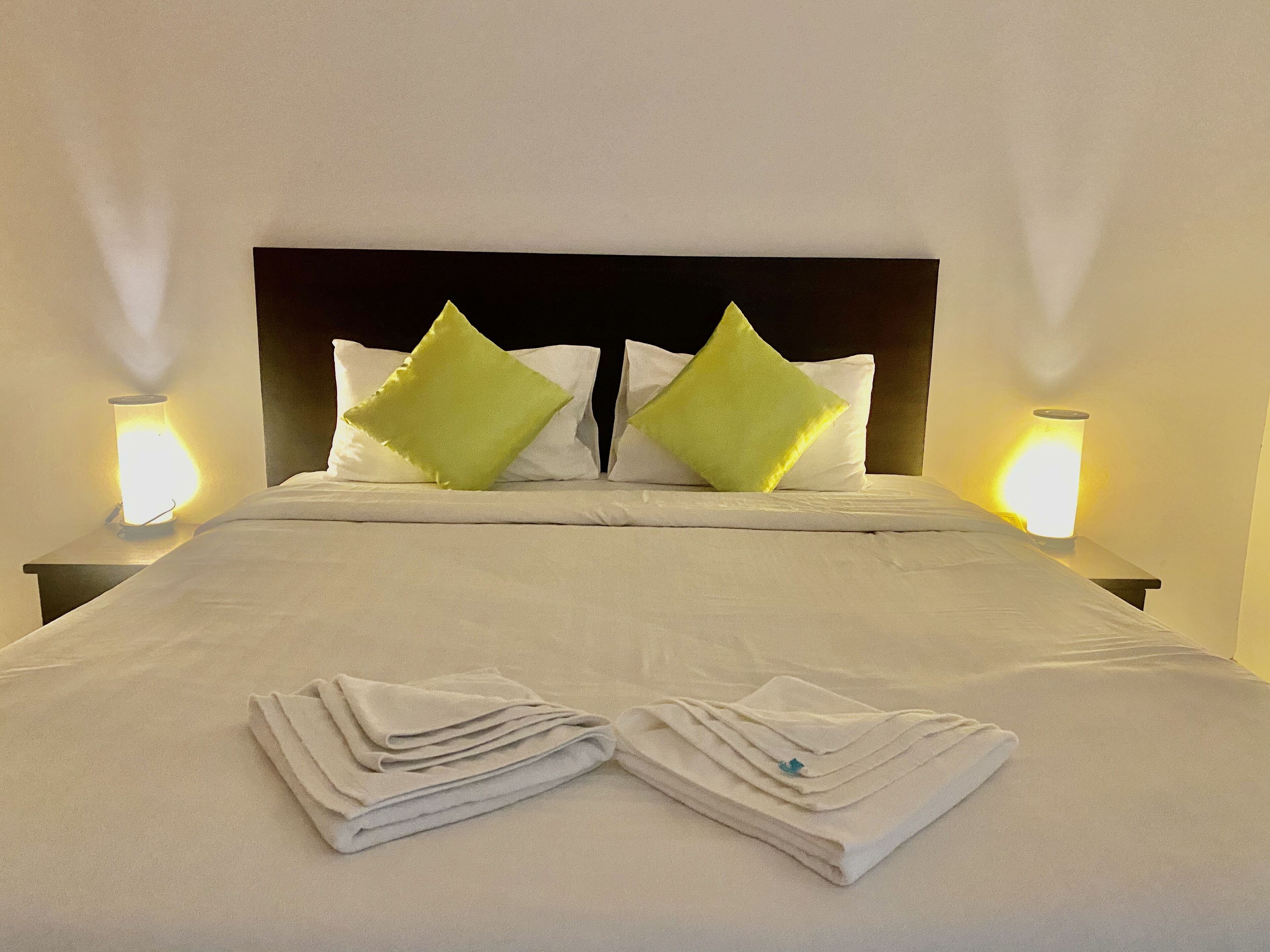 Una cama bien hecha con sábanas blancas y dos almohadas verdes, con dos lámparas de noche a cada lado.