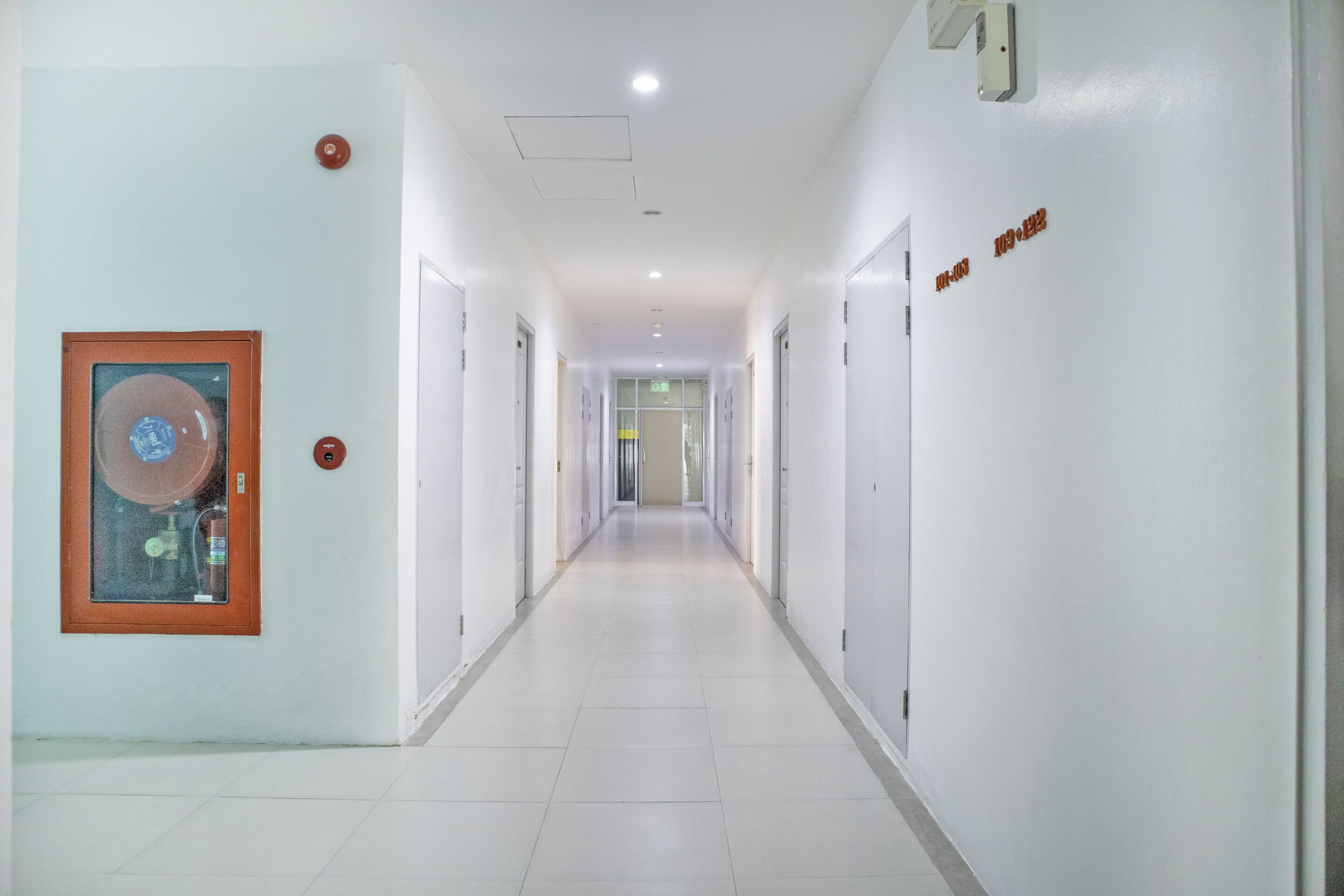 A long, narrow hallway with white walls and doors, a framed picture on the left, and a sign on the right.