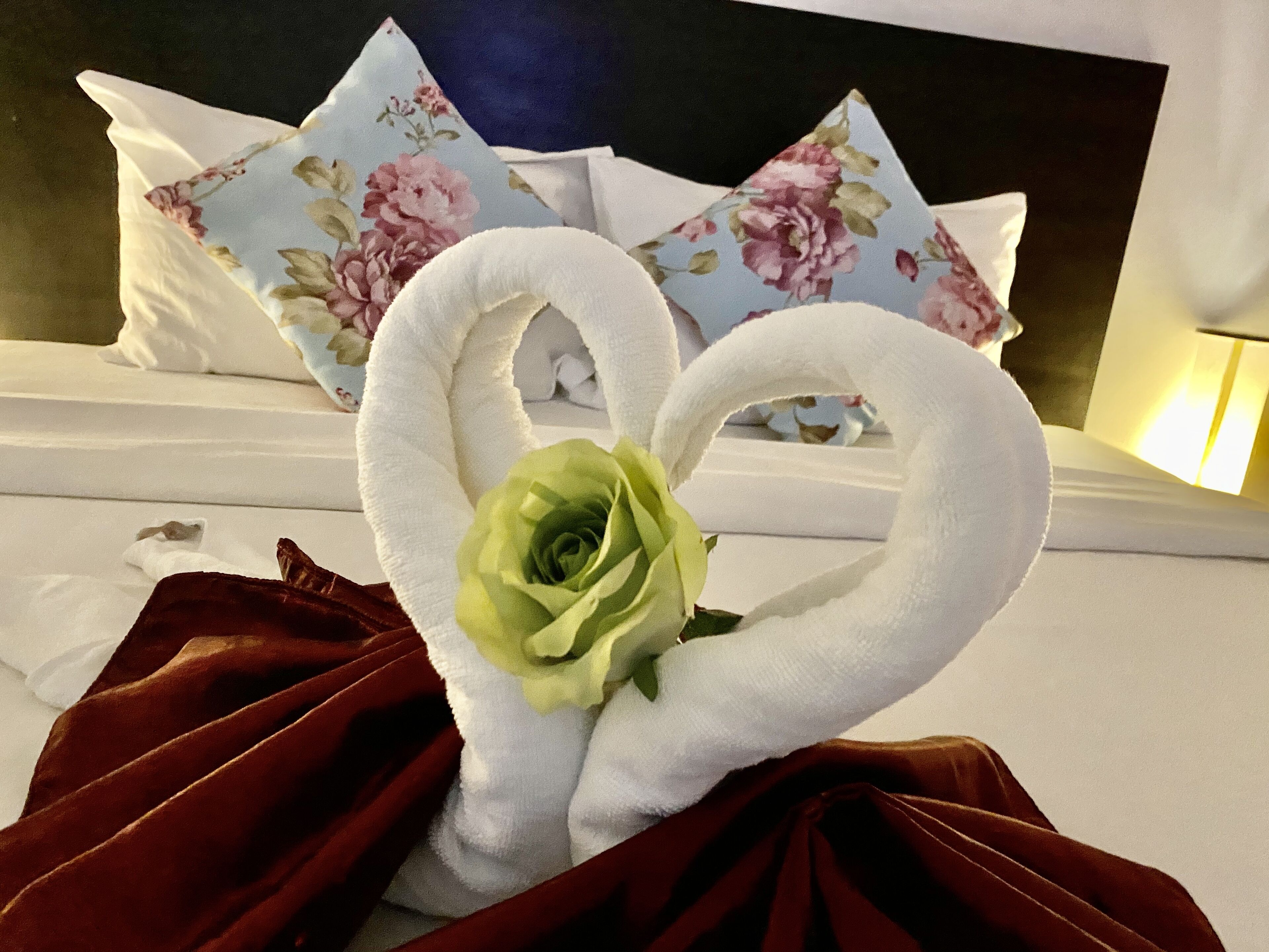 A bed with white bedding, a floral pillow, and a heart-shaped towel arrangement with a green rose.