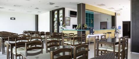 Un comedor amplio con varias mesas y sillas, una barra con dispensador de agua azul y un televisor montado en la pared.