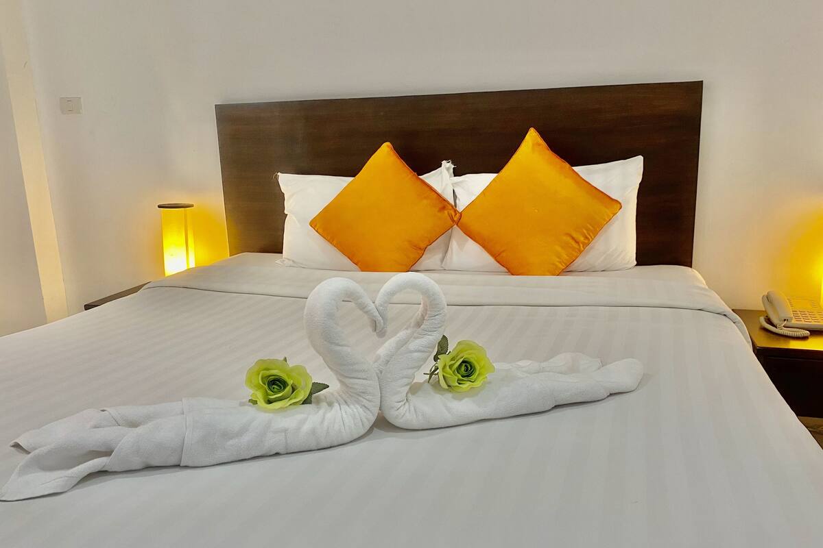 Una habitación de hotel con una cama bien hecha, dos almohadas anaranjadas, un cabecero blanco, una lámpara de noche y un teléfono.