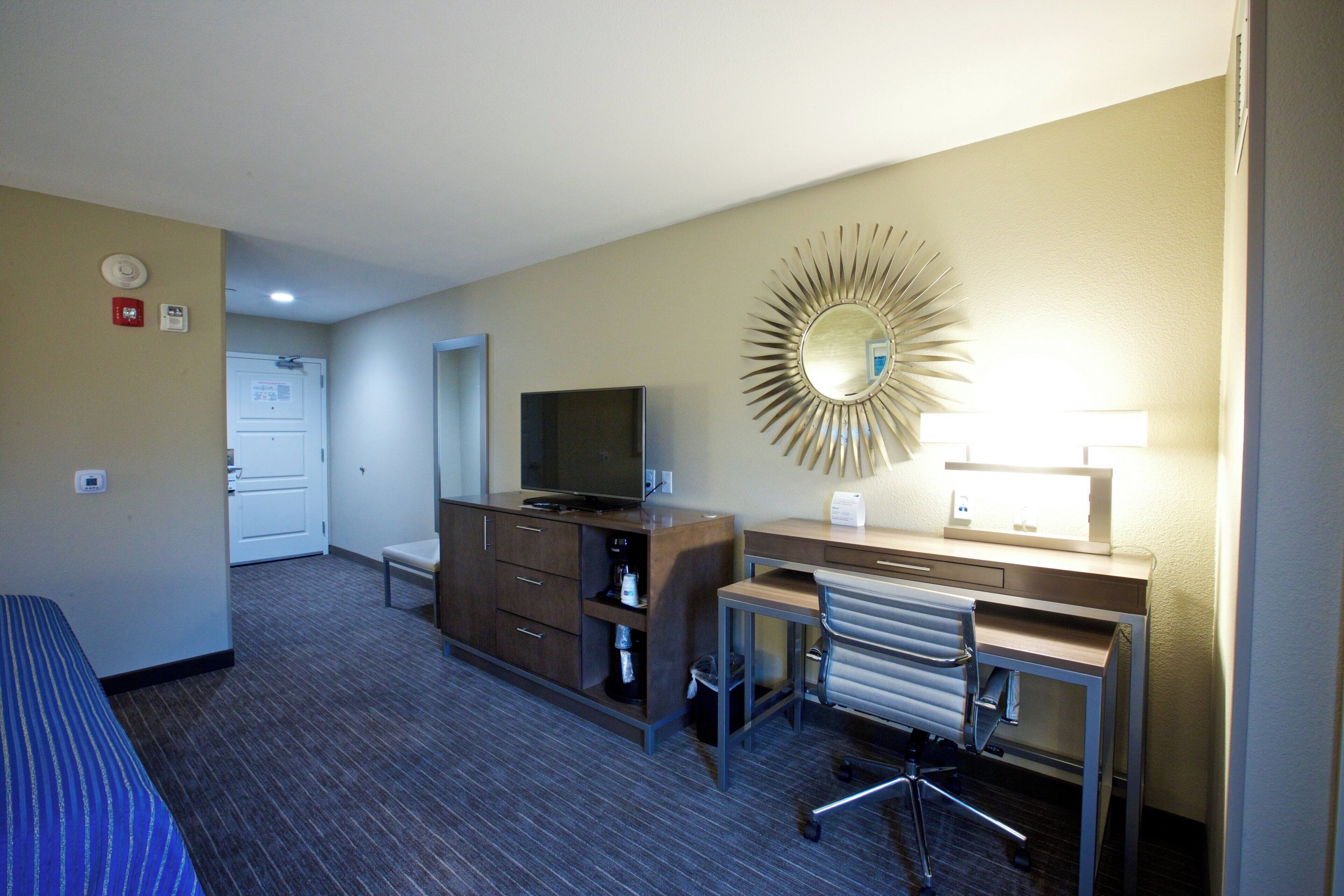 A hotel room with a bed, a desk, a chair, a television, a mirror, and a wall-mounted light.