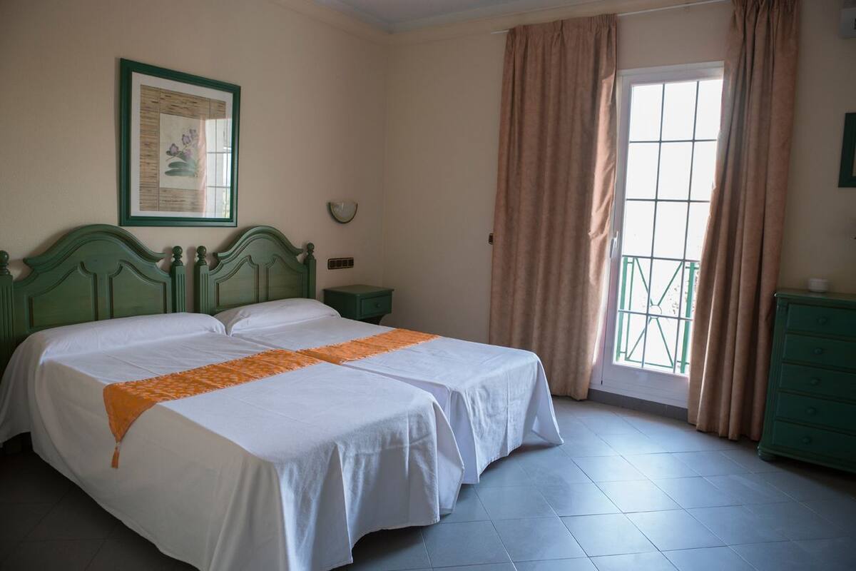 A double bed with white linens and an orange throw, a green headboard, a framed picture on the wall, a nightstand, and a window with curtains.