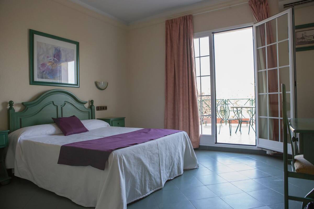 A bedroom with a bed, a green headboard, a purple pillow, a white bedspread, a green nightstand, a framed picture on the wall, and a balcony with a table and chairs.
