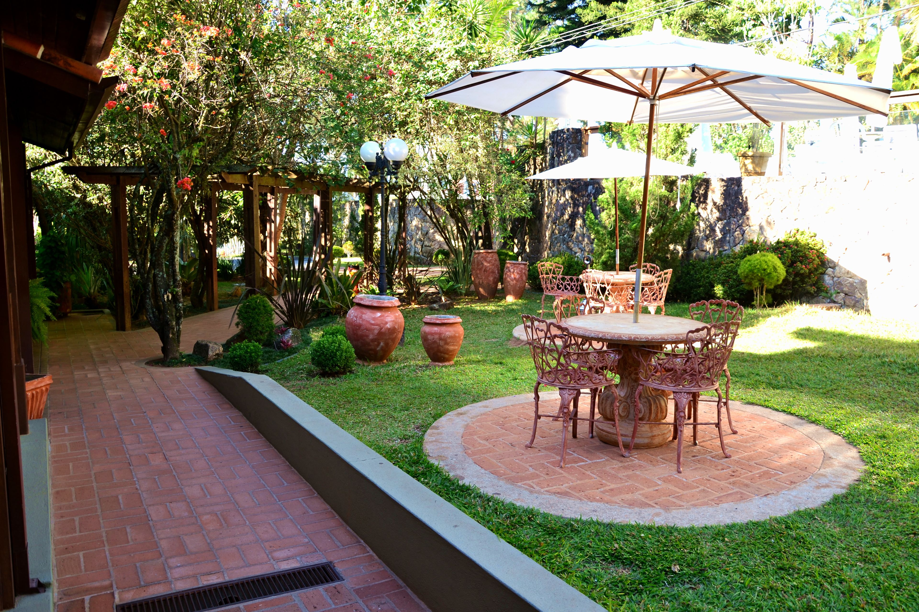 A patio area with a brick floor, a round table with chairs, and an umbrella. There are potted plants and a pergola with hanging lights.