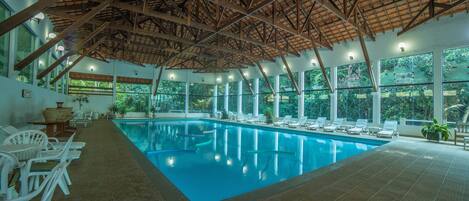 An indoor swimming pool with clear blue water, surrounded by white lounge chairs and tables, large windows offering views of greenery, and a wooden ceiling structure.
