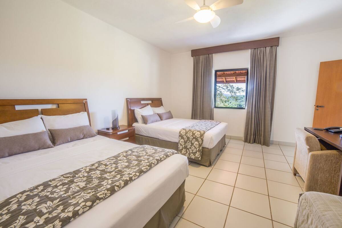 A hotel room with two beds, a desk, a chair, a ceiling fan, and a window with curtains.