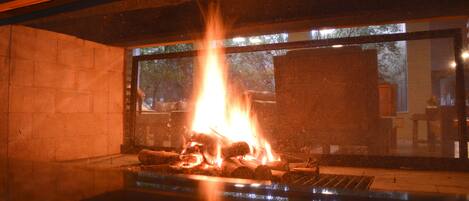 Una estructura con fuego, leña y una parrilla, una pared de piedra y una ventana con vista a árboles.