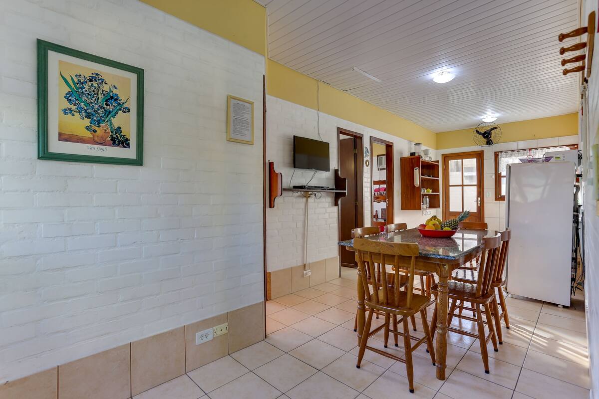 A dining area with a wooden table and chairs, a television mounted on the wall, and a refrigerator in the background.
