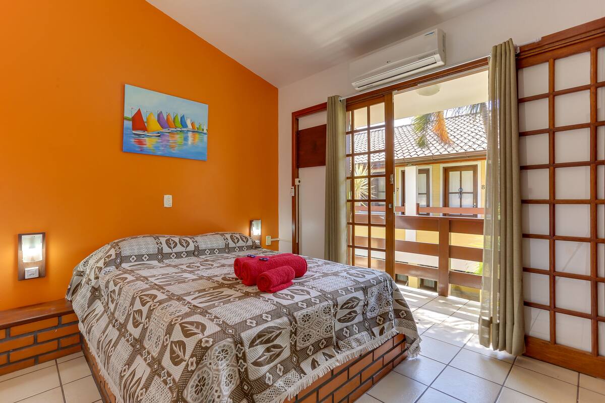 A bedroom with a large bed, a painting of sailboats, an air conditioner, and a view through a sliding door.