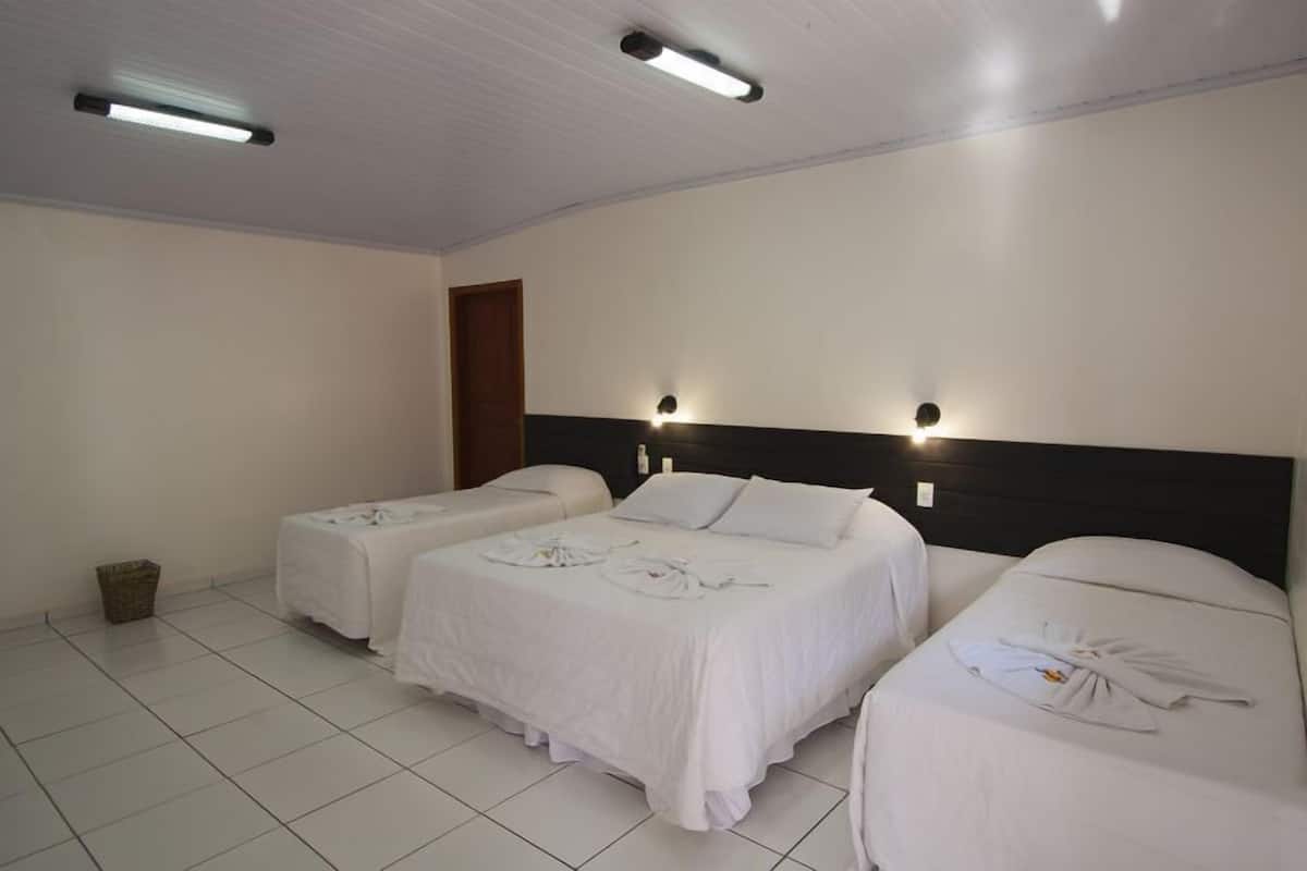 Ein Hotelzimmer mit zwei Einzelbetten und einem größeren Bett, alle mit weißer Bettwäsche. Über jedem Bett befinden sich zwei Wandleuchten und an der Decke eine einzelne Leuchte.