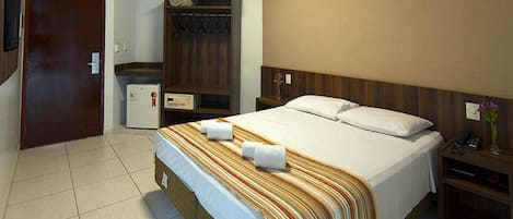 A hotel room with a large bed, a wardrobe, a small fridge, and a bedside table with a lamp.