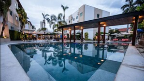 A swimming pool with a covered area, palm trees, and buildings in the background. - Hotel Morada do Sol (Caldas Novas)