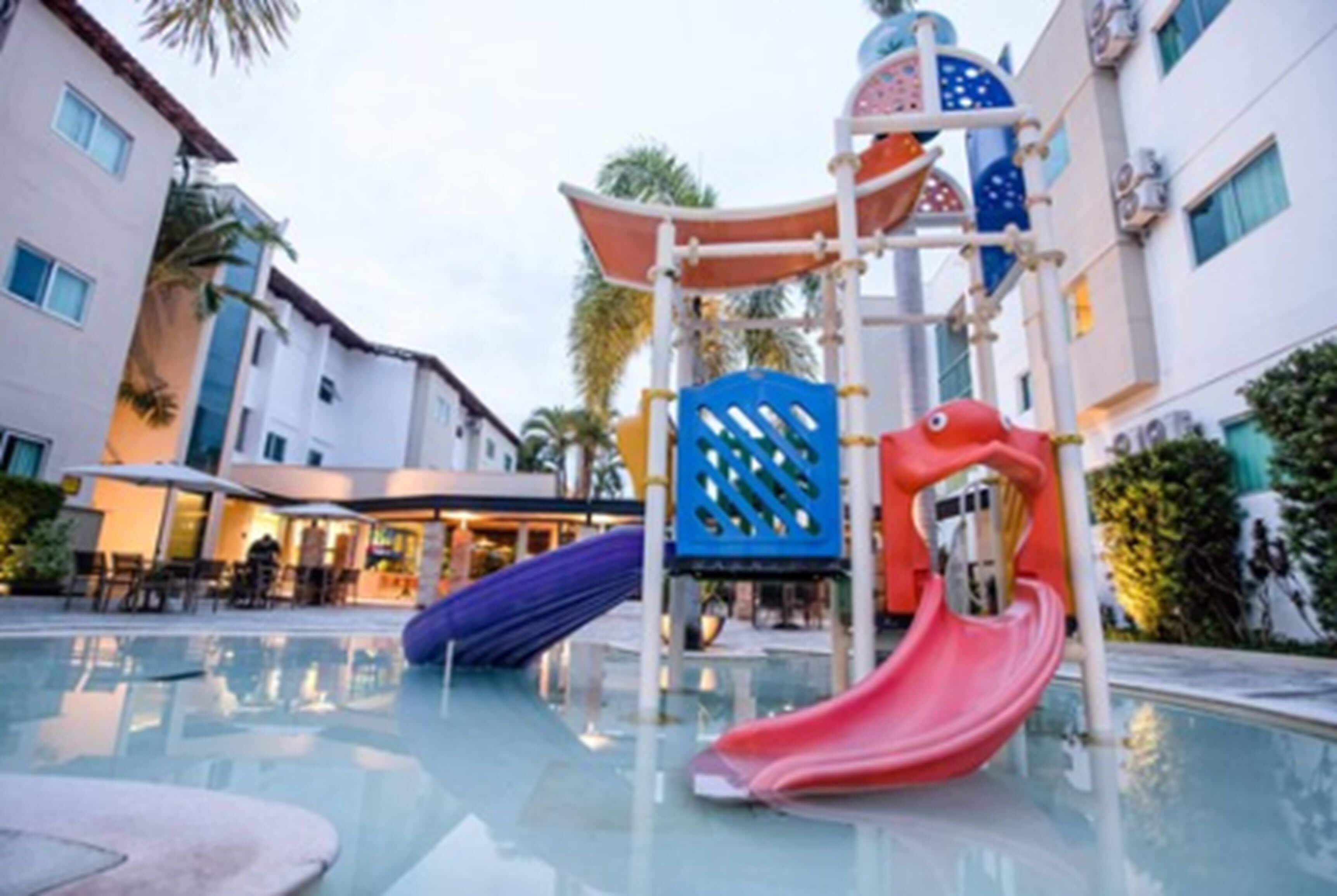 Zona infantil con toboganes y estructuras para escalar, rodeada por una piscina y edificios de hotel.