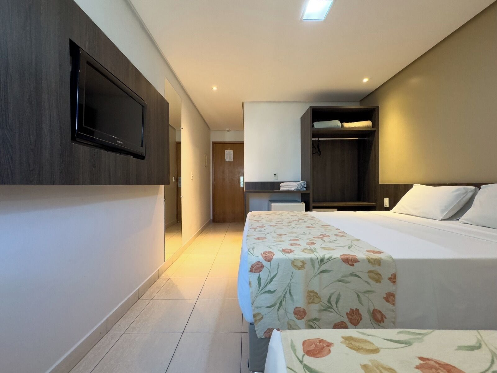 A hotel room with a bed, a TV mounted on the wall, a wardrobe, and a small desk with a chair.
