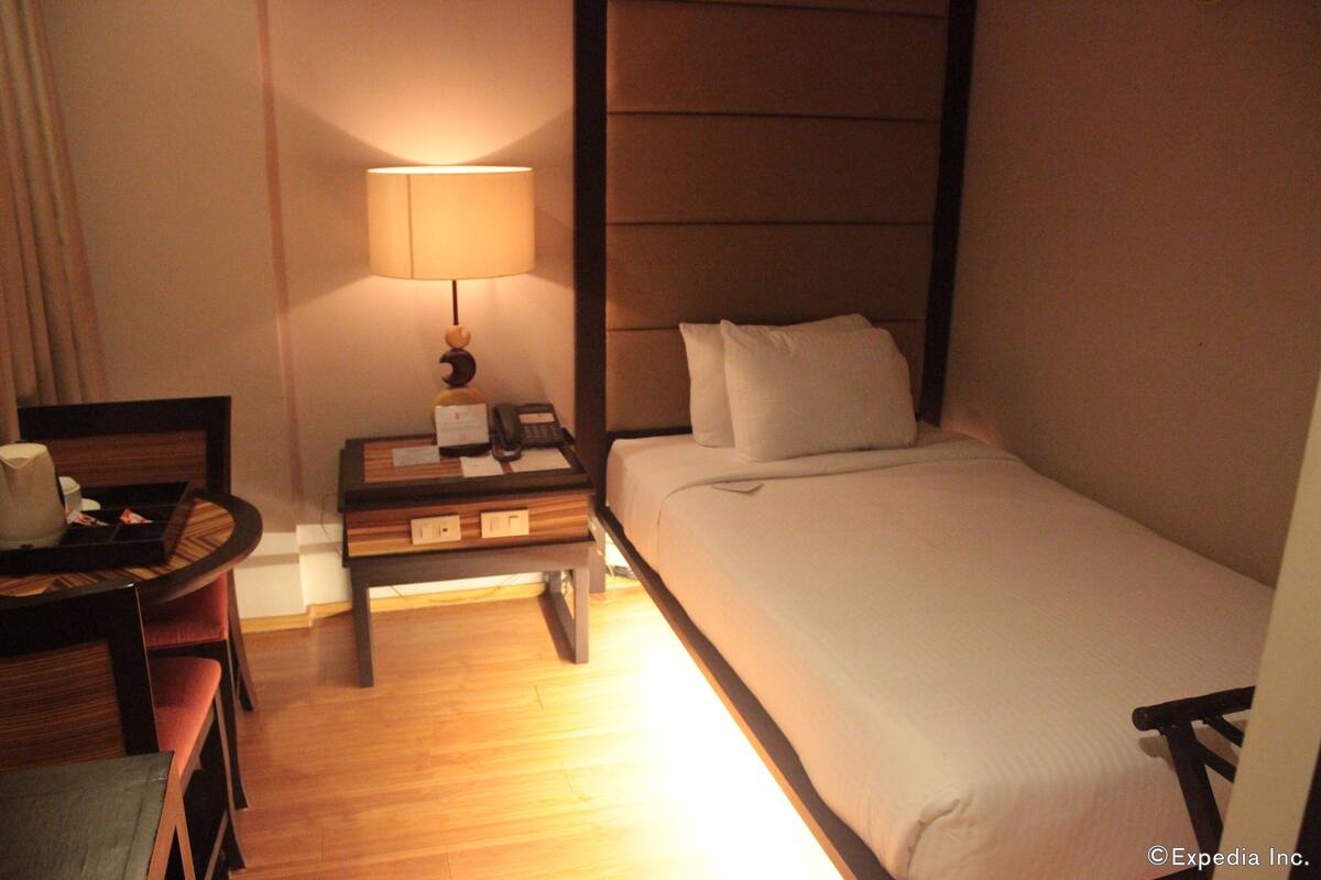 Quarto de hotel com uma cama, mesa de cabeceira com abajur, uma cadeira e escrivaninha com telefone.