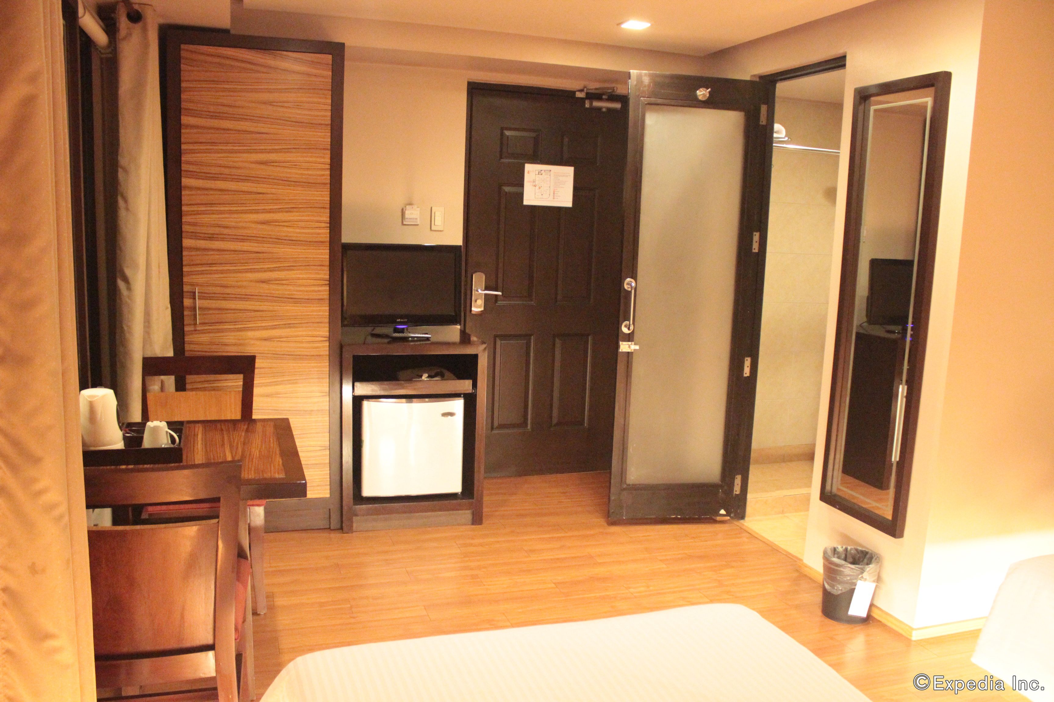 A hotel room with a bed, a desk, a chair, a small refrigerator, a trash bin, and a mirror.