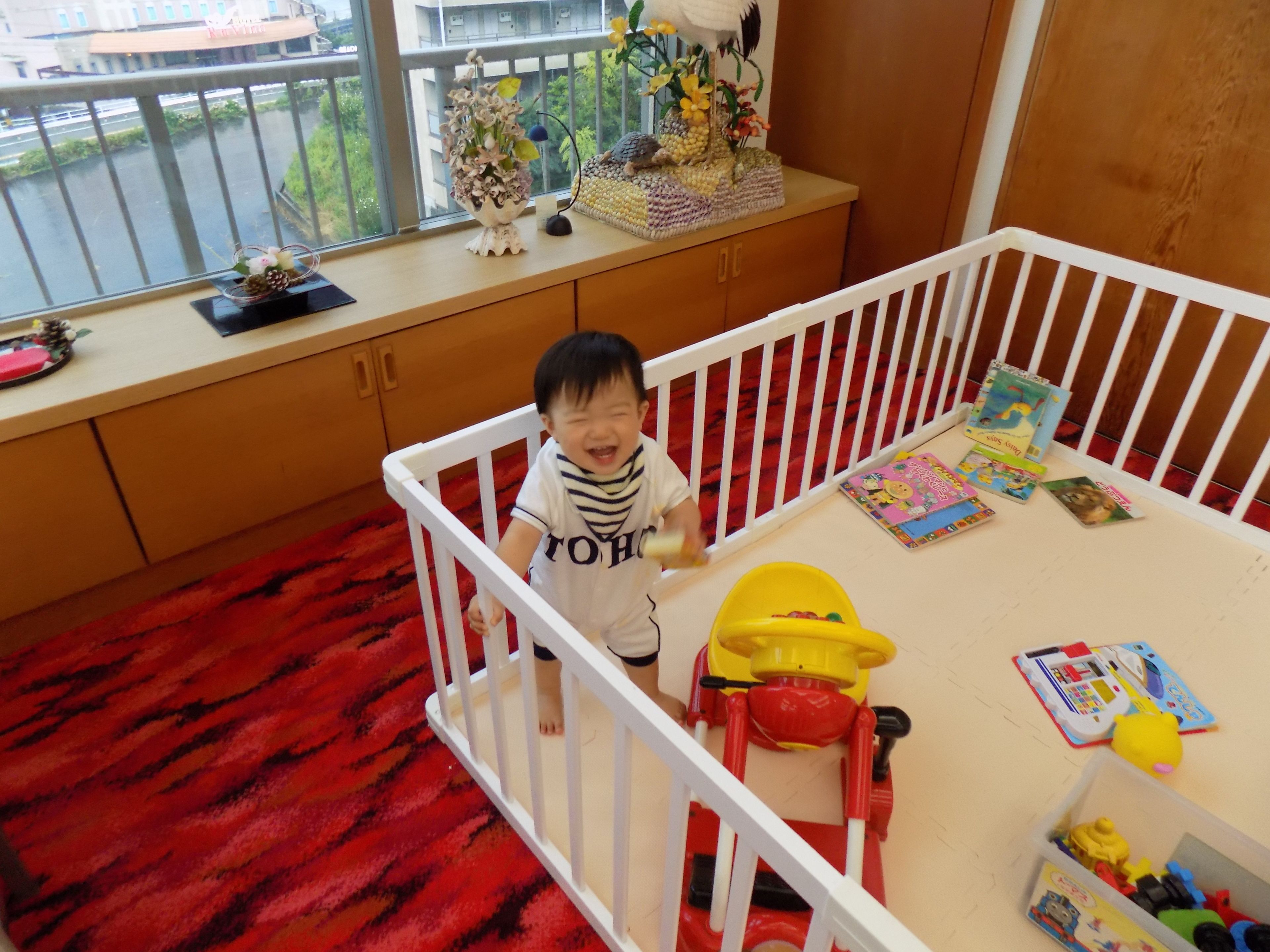 A child in a white crib with a red and black patterned carpet, a white railing, and a window with a view of buildings and greenery.