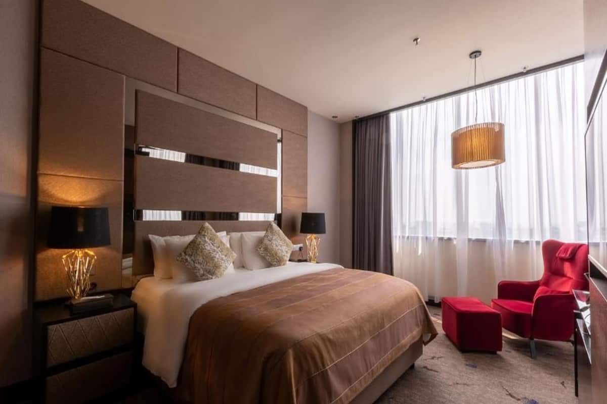A modern hotel room with a large bed, a red armchair, and a large window with curtains.