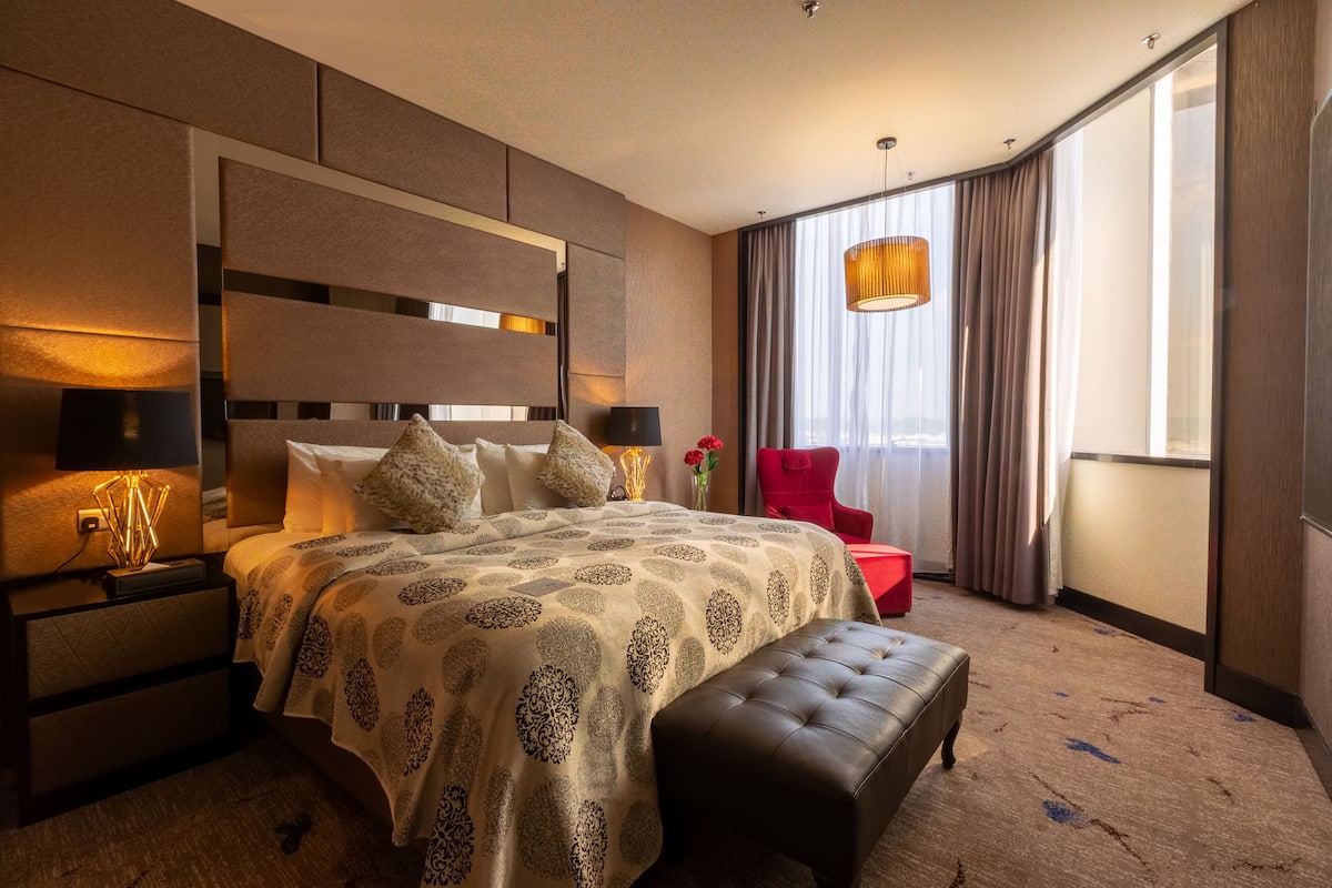 A hotel room with a large bed, a red chair, a bench, a nightstand, and a large window with curtains.