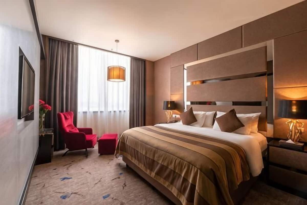 A hotel room with a large bed, a red armchair, a small table, and a large window with curtains.