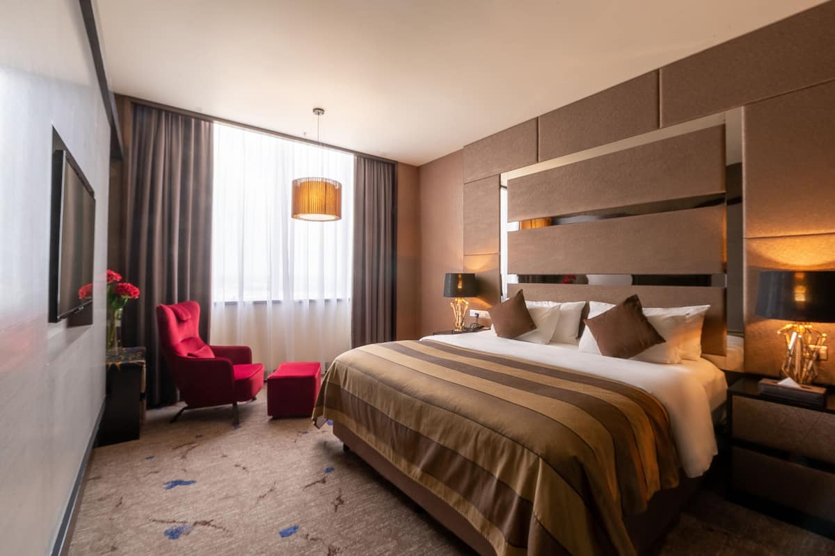 A hotel room with a large bed, a red armchair, a small table, and a large window with curtains.