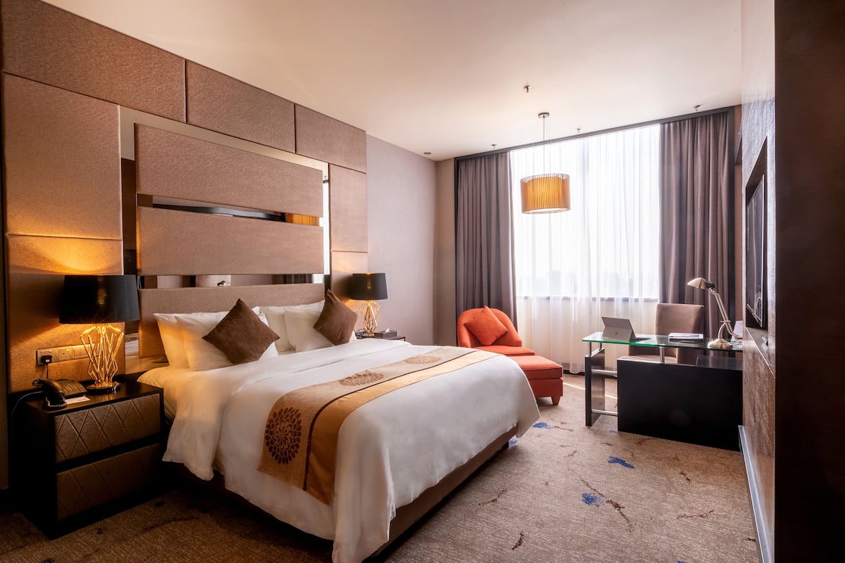 A hotel room with a large bed, a desk with a laptop, a chair, a nightstand with a lamp, and a large window with curtains.