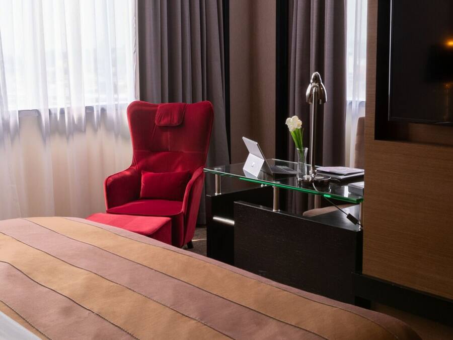 Une chambre d’hôtel avec un lit, un fauteuil rouge, un bureau en verre avec un ordinateur portable, un vase avec des fleurs et un téléviseur fixé au mur.