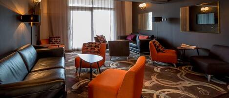 Une chambre d’hôtel moderne avec un canapé en cuir, des fauteuils orange, une petite table et un tapis à motifs.