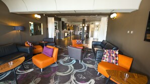 Un moderno vestíbulo de hotel con una variedad de zonas de estar, incluyendo sofás naranja y grises, un bar y una recepción.