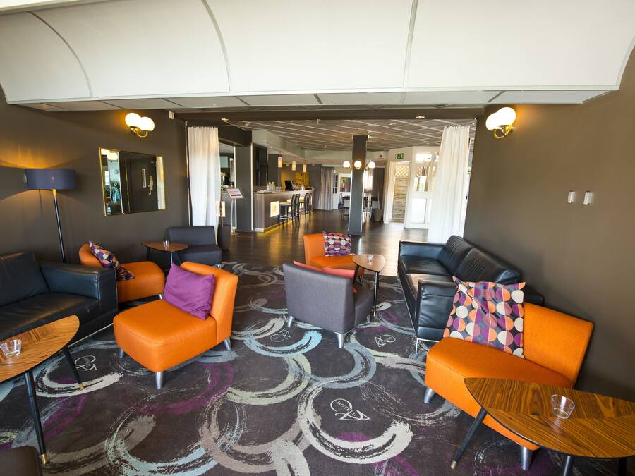 A modern hotel lobby with a mix of seating areas, including orange and gray sofas, a bar, and a reception desk.