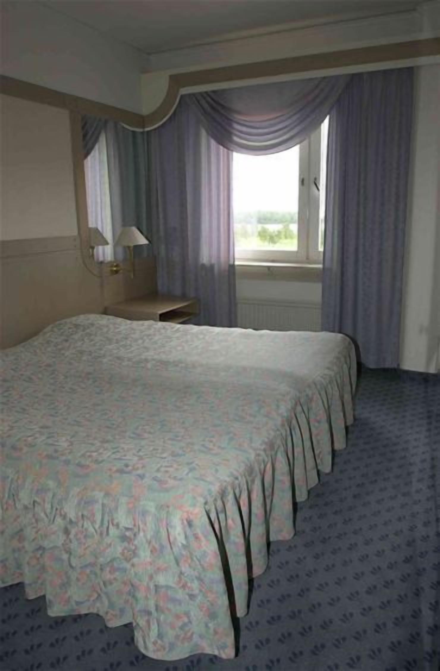 Een hotelkamer met een groot bed, een nachtkastje, een lamp en een raam met gordijnen.