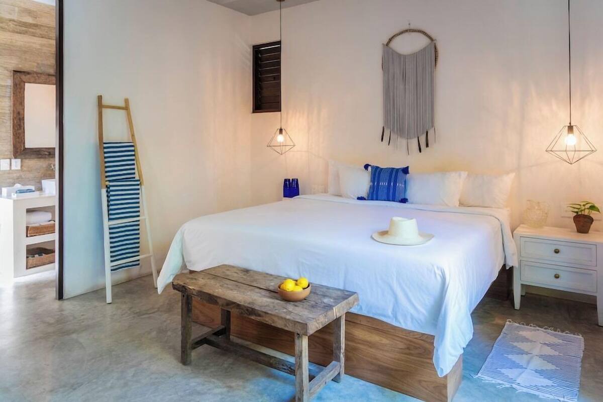 Une chambre à coucher comprenant un lit, des tables de chevet, un banc en bois, une étagère fixée au mur et un luminaire suspendu.