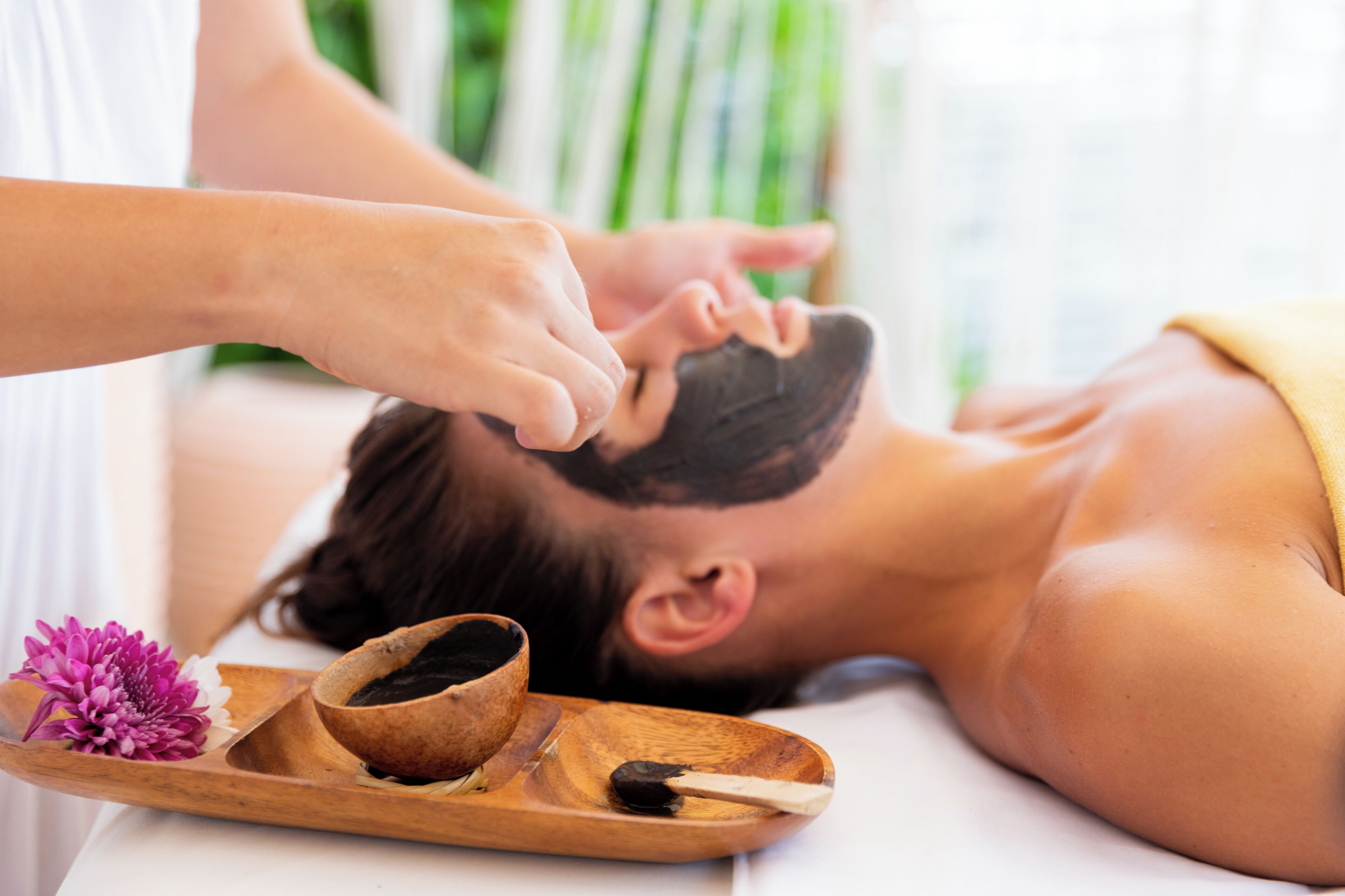 A person receiving a facial treatment with a wooden tray containing a bowl of black powder and a flower.