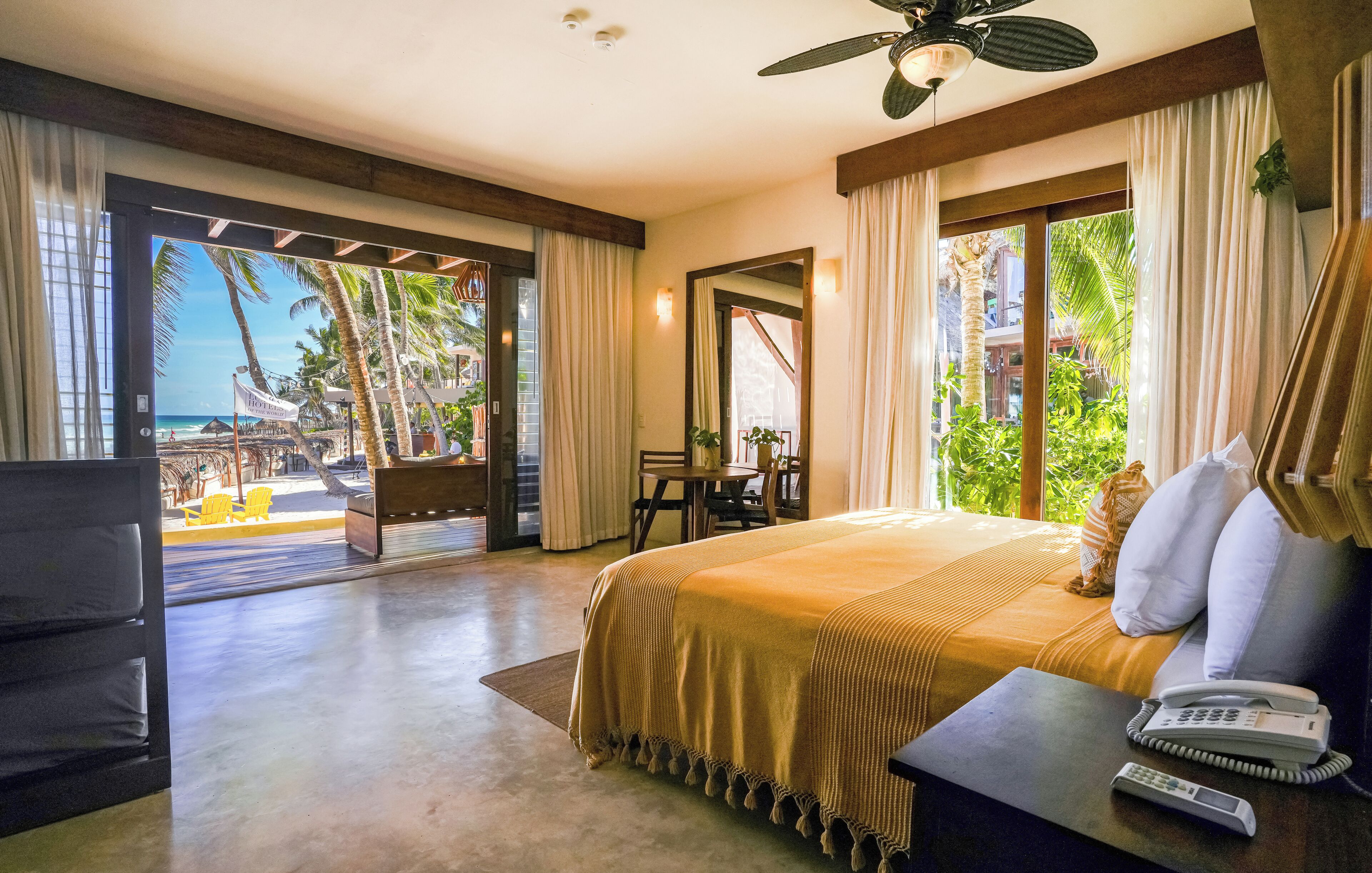 Een hotelkamer met een bed, een bureau met telefoon en uitzicht op een strand en palmbomen.