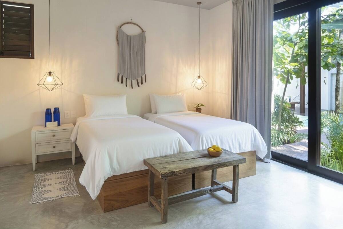 Une chambre avec deux lits, une table de chevet, une table en bois avec un bol, et une vue sur la verdure à l’extérieur.