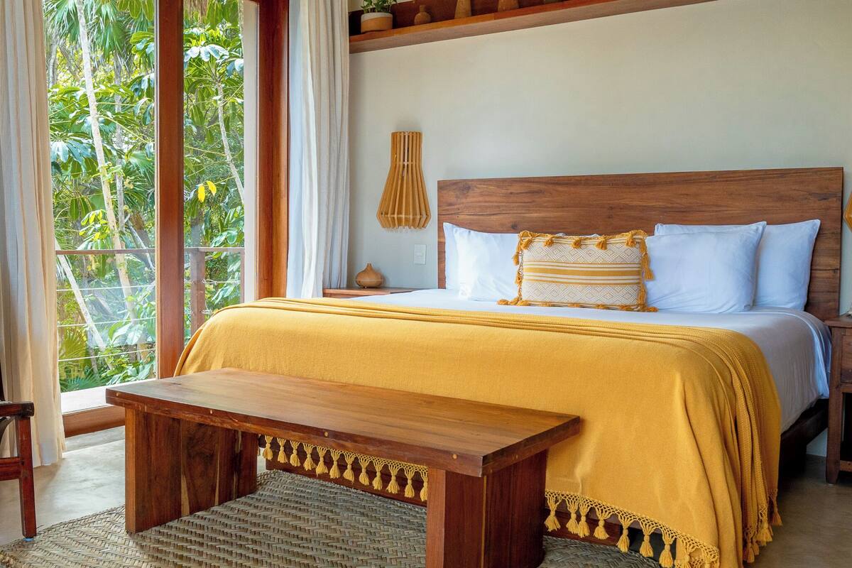 Une chambre avec un lit en bois, un banc, un ventilateur de plafond et une vue sur la verdure à l’extérieur.
