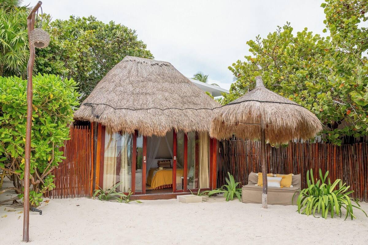 A beachside hut with a thatched roof, a bed with a yellow bedspread, and a small seating area with cushions.