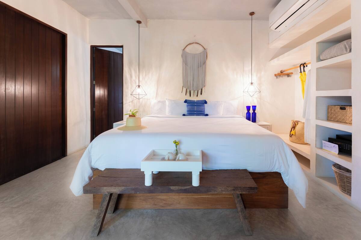Une chambre à coucher avec un grand lit, des tables de chevet, un banc en bois, une étagère fixée au mur et un luminaire suspendu.