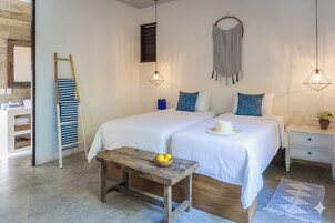 A bedroom with two beds, a wooden bench with a bowl of fruit, a bedside table with a plant, and a wall-mounted macrame piece.